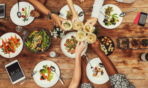 overhead view of a dining table with food and a close-up of a group of diverse women's hands holding wine glasses together. How To Enjoy Food.