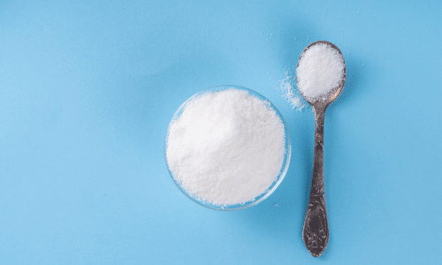 allulose powder in a glass bowl and a teaspoonful on a blue background.