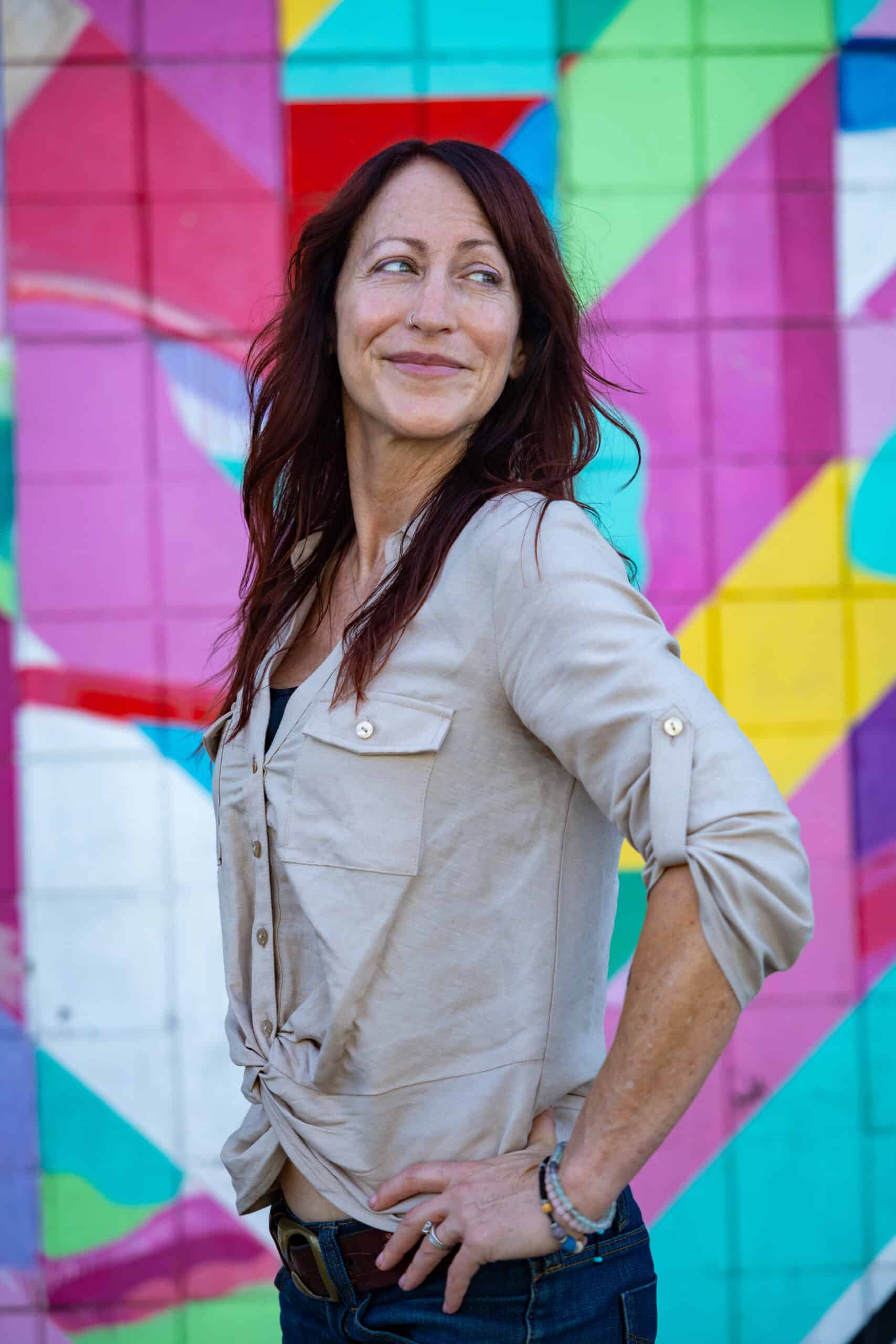Maria with hands on her hips looking over her shoulder in front of a brightly colored mural wall. Veg Out With Maria