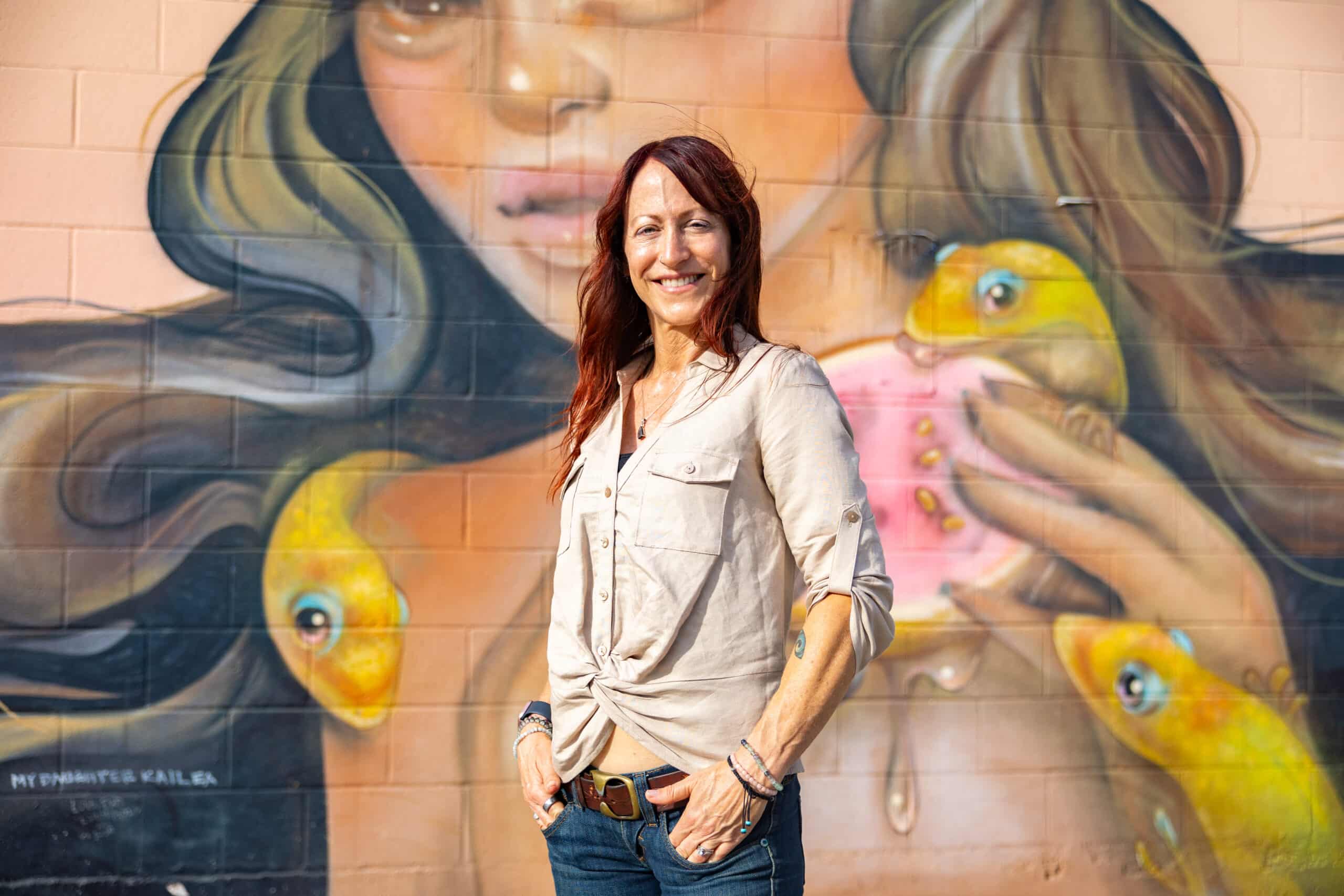 Maria with hands in pockets in front of a mural of a Hawaiian woman with geckos and guava fruit. Veg Out With Maria