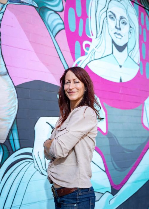 Maria Tointon Maria with arms crossed in front of a mural of a woman. Veg Out With Maria